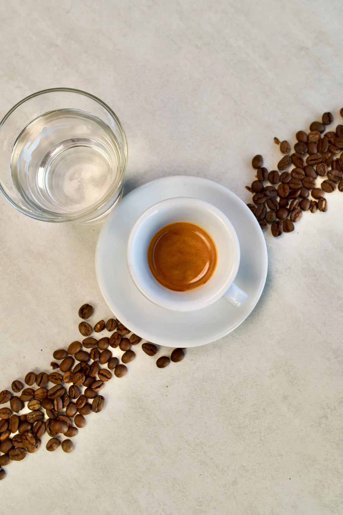 Espresso shot in a white cup with coffee beans and a glass of water on a light surface