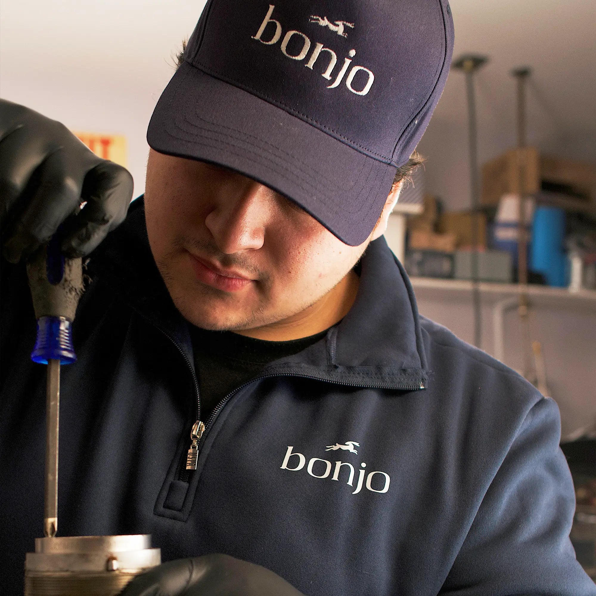 A person wearing a navy cap and jacket with "bonjo" logo is focused on using a screwdriver. They are in a workshop, showing concentration and precision.