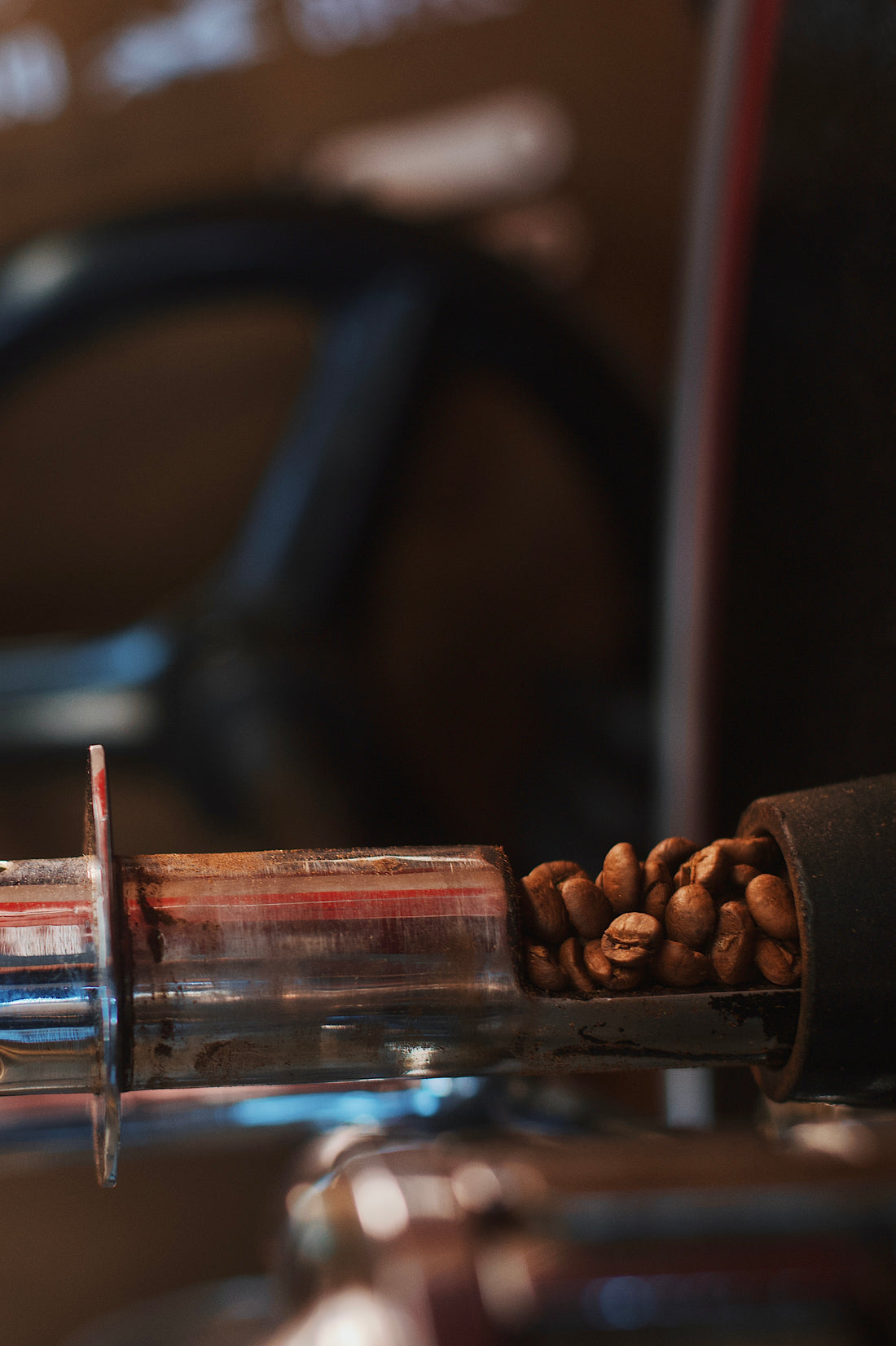 Close-up of coffee beans in a metal roasting tube, with a blurred background. The scene conveys warmth and the artisanal process of coffee making.