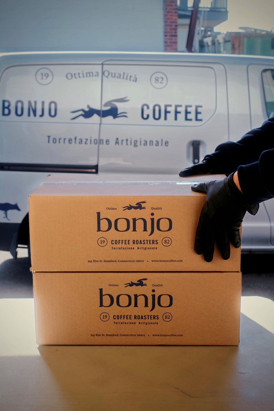 Person wearing a black glove reaching for a 'bonjo coffee' box in front of a van with the same brand logo.